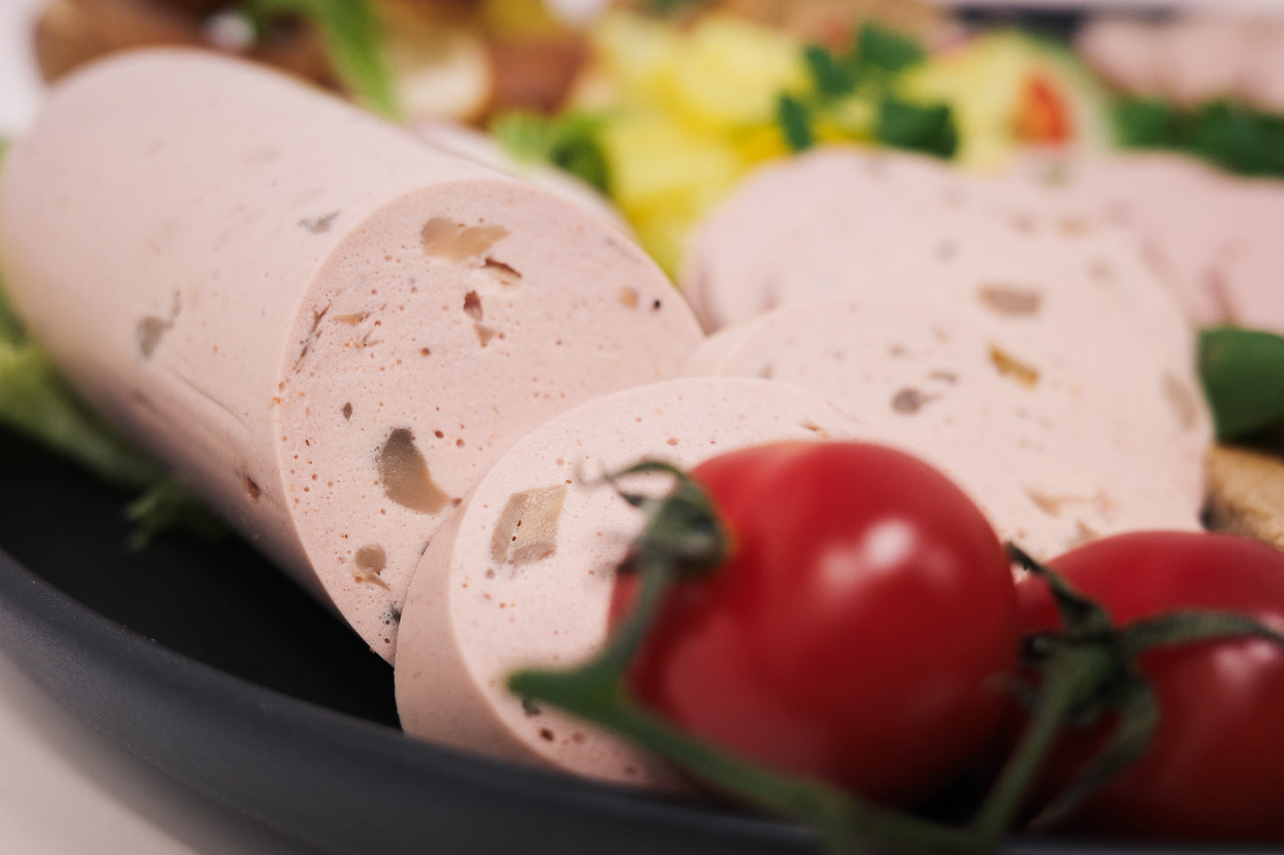 Veganer Champignon Lyoner zum Teil aufgeschnitten mit mehreren Tomaten im Vordergrund.